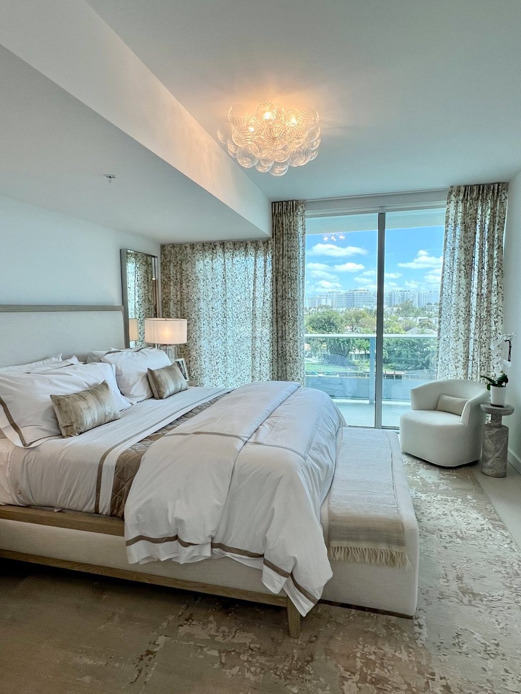 Bedroom with a glass bubble ceiling fixture, layered white bedding, patterned drapery and a balcony view over the water