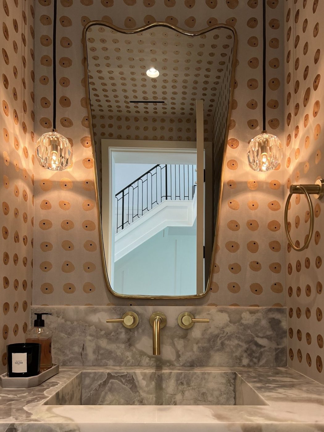 Powder room with hand-painted dot wallpaper, a wavy brass mirror, faceted crystal pendants, brass wall-mounted faucet and an onyx sink